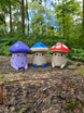 Three Palia Fun Guy 3D printed figurines with purple, blue, and red mushroom caps displayed on a tree stump in a forest.