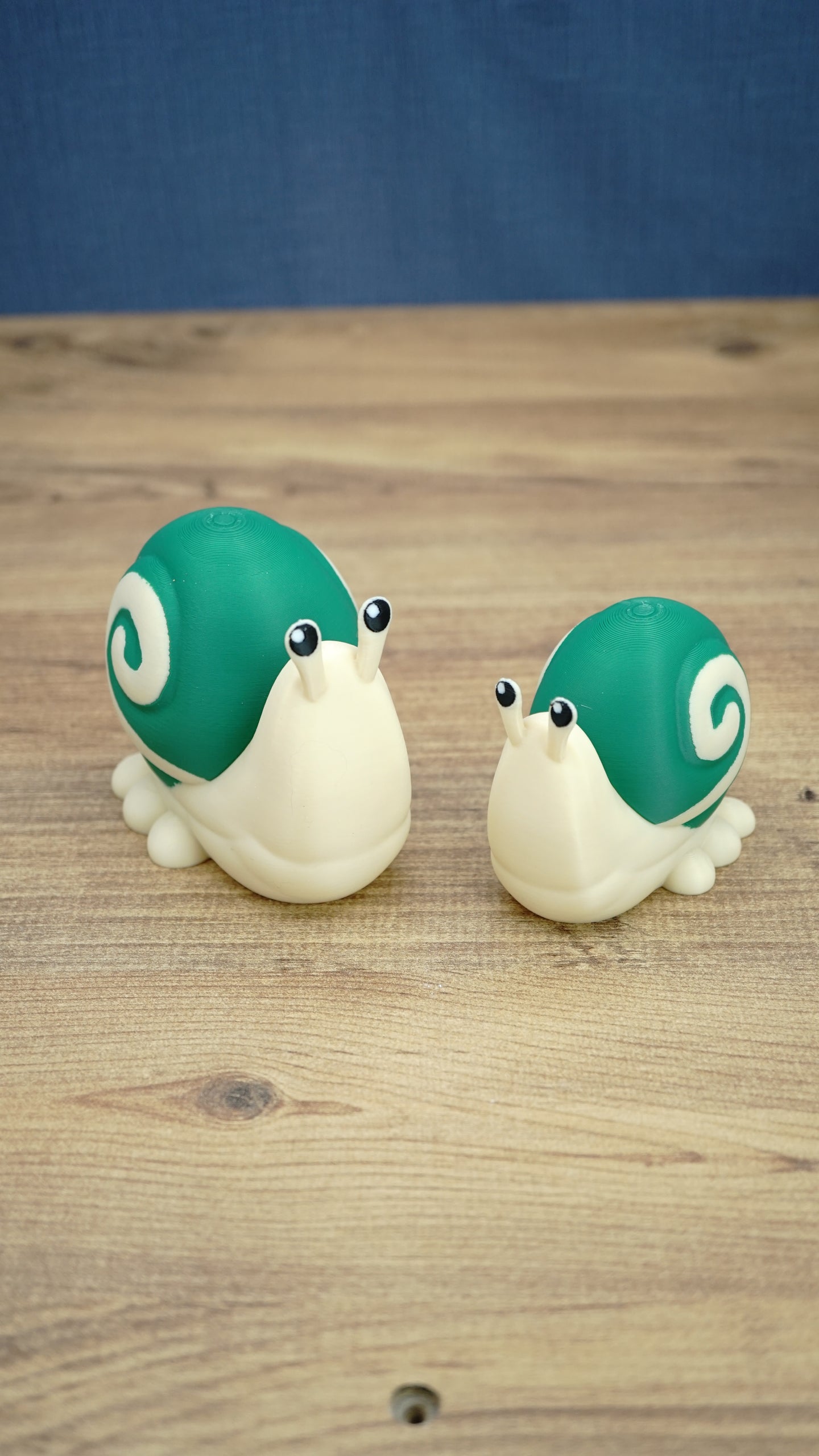 Palia Garden Snail Plush collectible figures in 8cm and 10cm sizes, featuring cream bodies, green spiral shells and cute raised eyes on a wooden surface