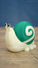 Side view of a Palia Garden Snail Plush figure showing its rounded green shell, cream spiral pattern, cute eye stalks and small snail feet