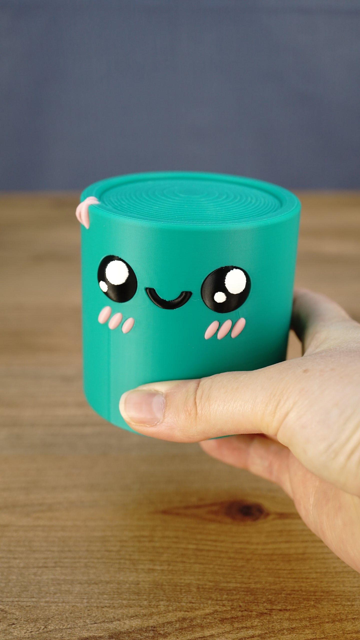 Close-up of a teal Palia Green Cylinder plush being held, showing its smiling face, black eyes, pink cheeks, and 3D printed texture.