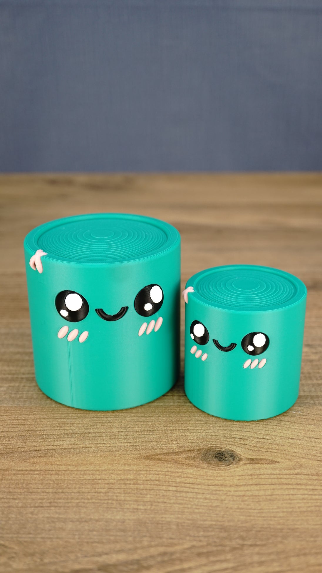 Two teal 3D printed Palia Green Cylinder plush figures in large and small sizes displayed together on a wooden tabletop.