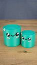 Two teal 3D printed Palia Green Cylinder plush figures in large and small sizes displayed together on a wooden tabletop.