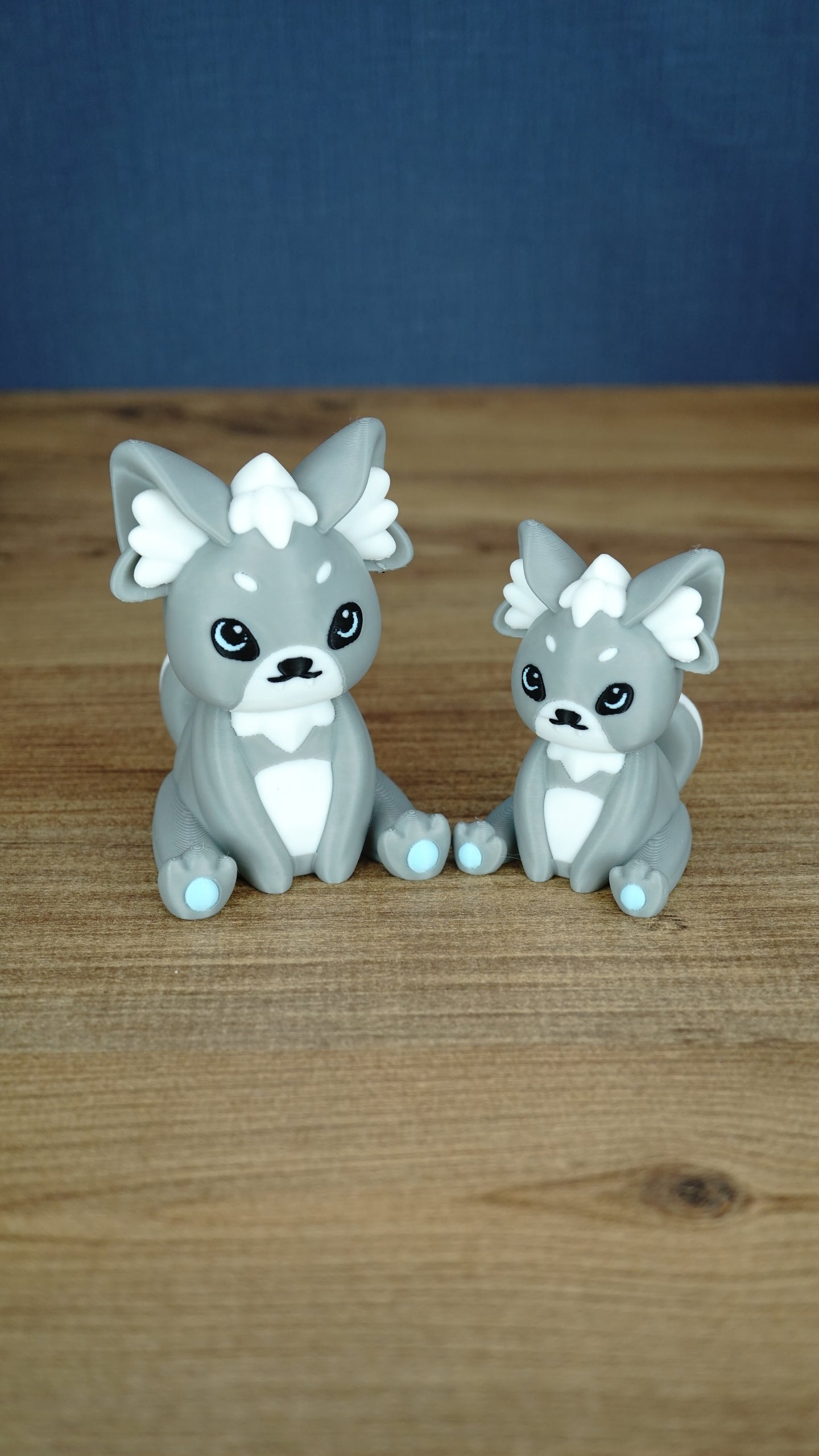 Two grey 3D printed Palcat plush figures with white accents, black eyes, light blue paw pads, and curled tails on a wooden tabletop.