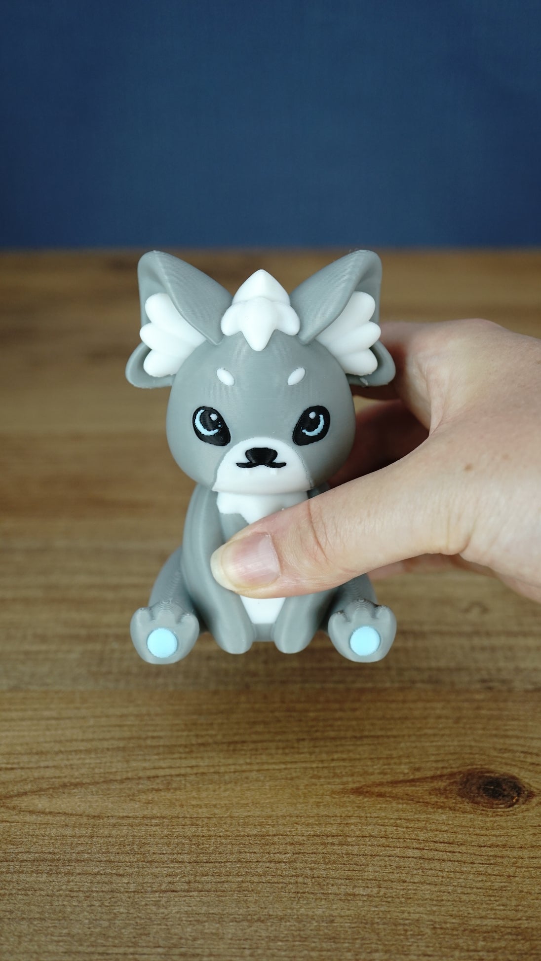 Handheld close-up of a grey 3D printed Palcat plush figure with white ear tufts, black eyes, white muzzle, and light blue paw pads.