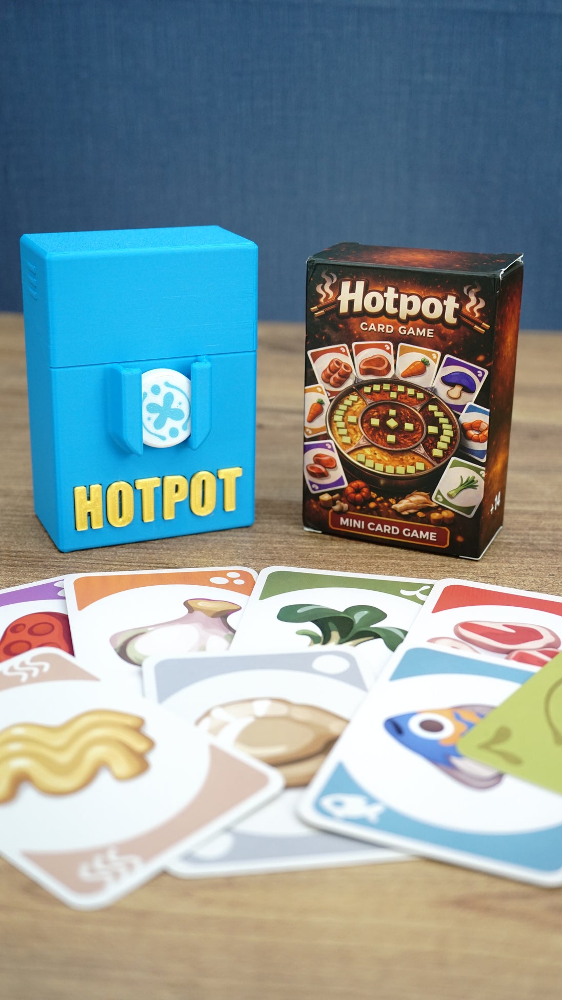 Palia Hotpot mini card game box and blue storage case shown behind a spread of colourful ingredient cards on a wooden tabletop.