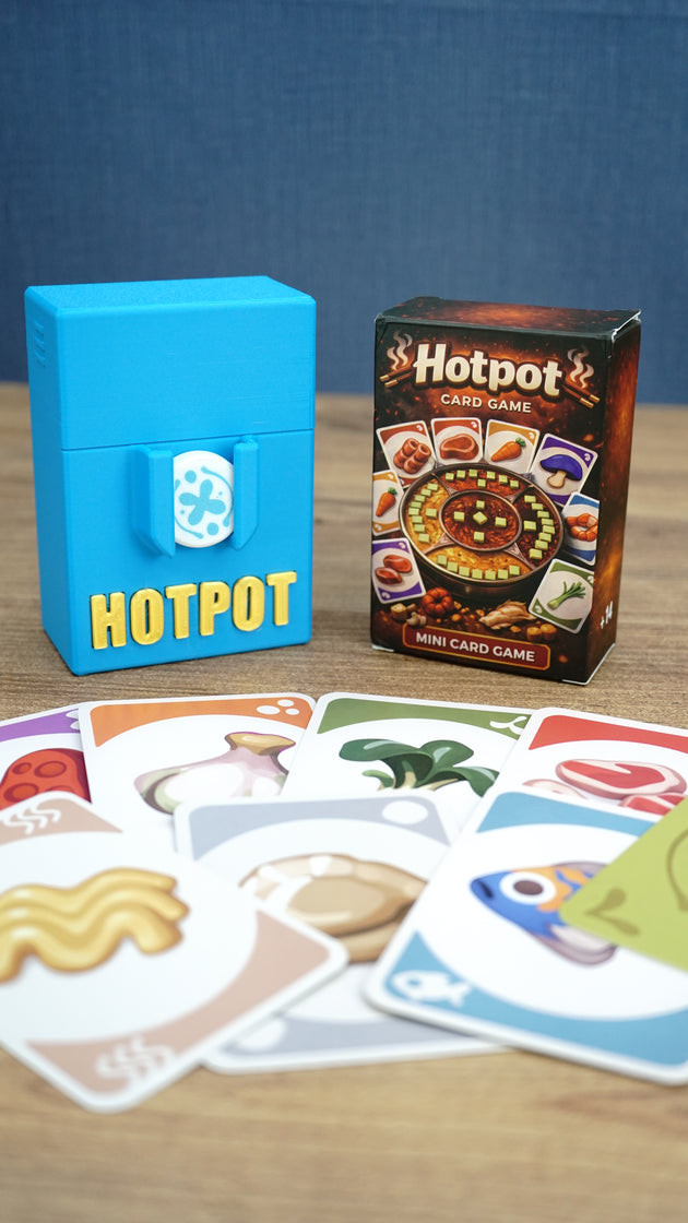 Palia Hotpot mini card game box and blue storage case shown behind a spread of colourful ingredient cards on a wooden tabletop.