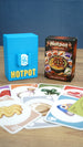 Palia Hotpot mini card game box and blue storage case shown behind a spread of colourful ingredient cards on a wooden tabletop.