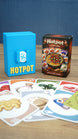 Palia Hotpot mini card game box and blue storage case shown behind a spread of colourful ingredient cards on a wooden tabletop.