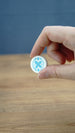 Close-up of a hand holding the white and blue Palia Hotpot pass token against a wooden tabletop background.