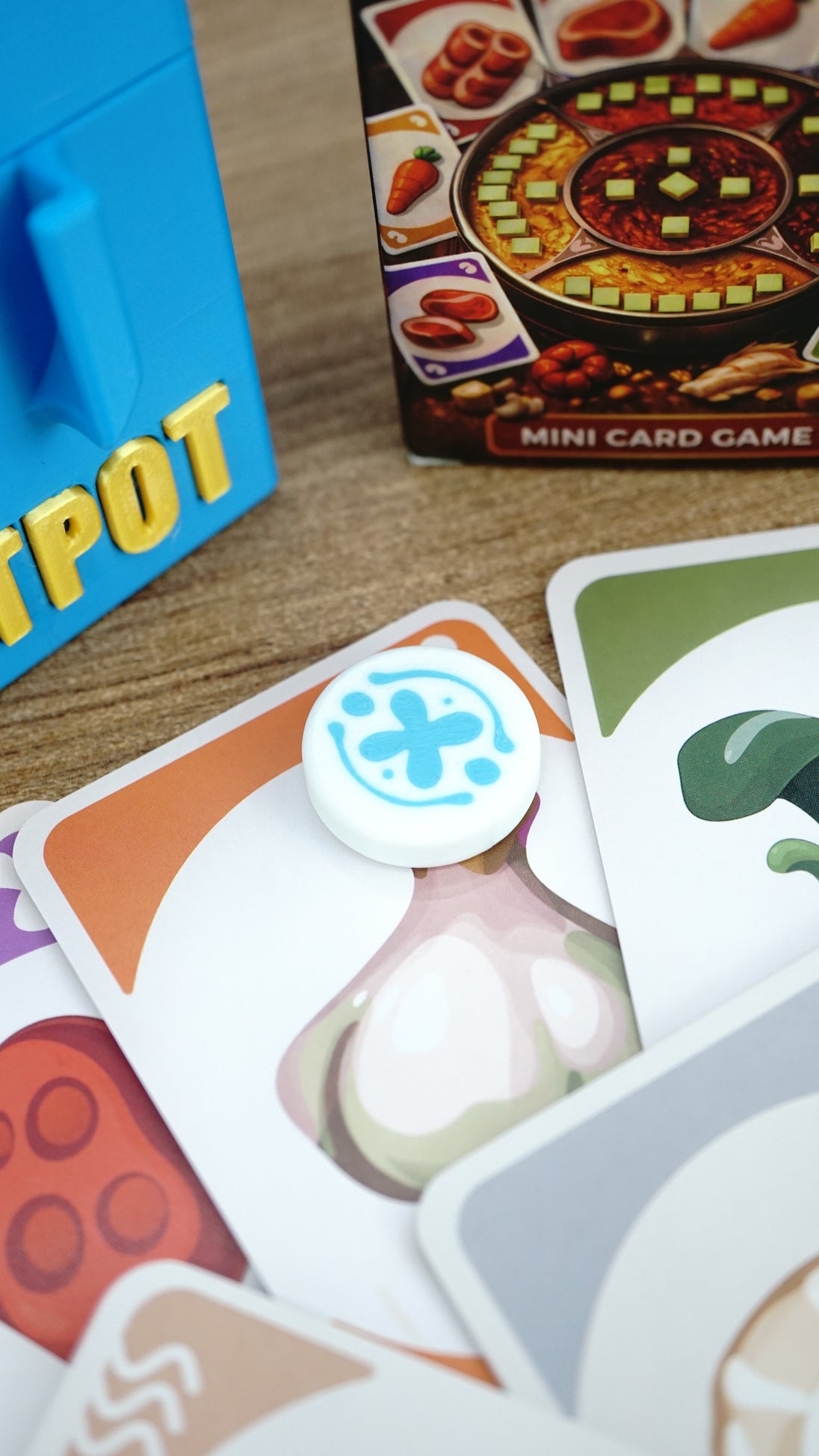 Close-up of a white and blue Palia Hotpot pass token resting on ingredient cards, with the card game box and blue storage case in the background.