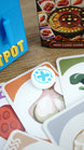 Close-up of a white and blue Palia Hotpot pass token resting on ingredient cards, with the card game box and blue storage case in the background.