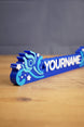 Close side view of a blue custom Palia-style nameplate showing raised white lettering, layered swirl accents, and star details for gamer decor