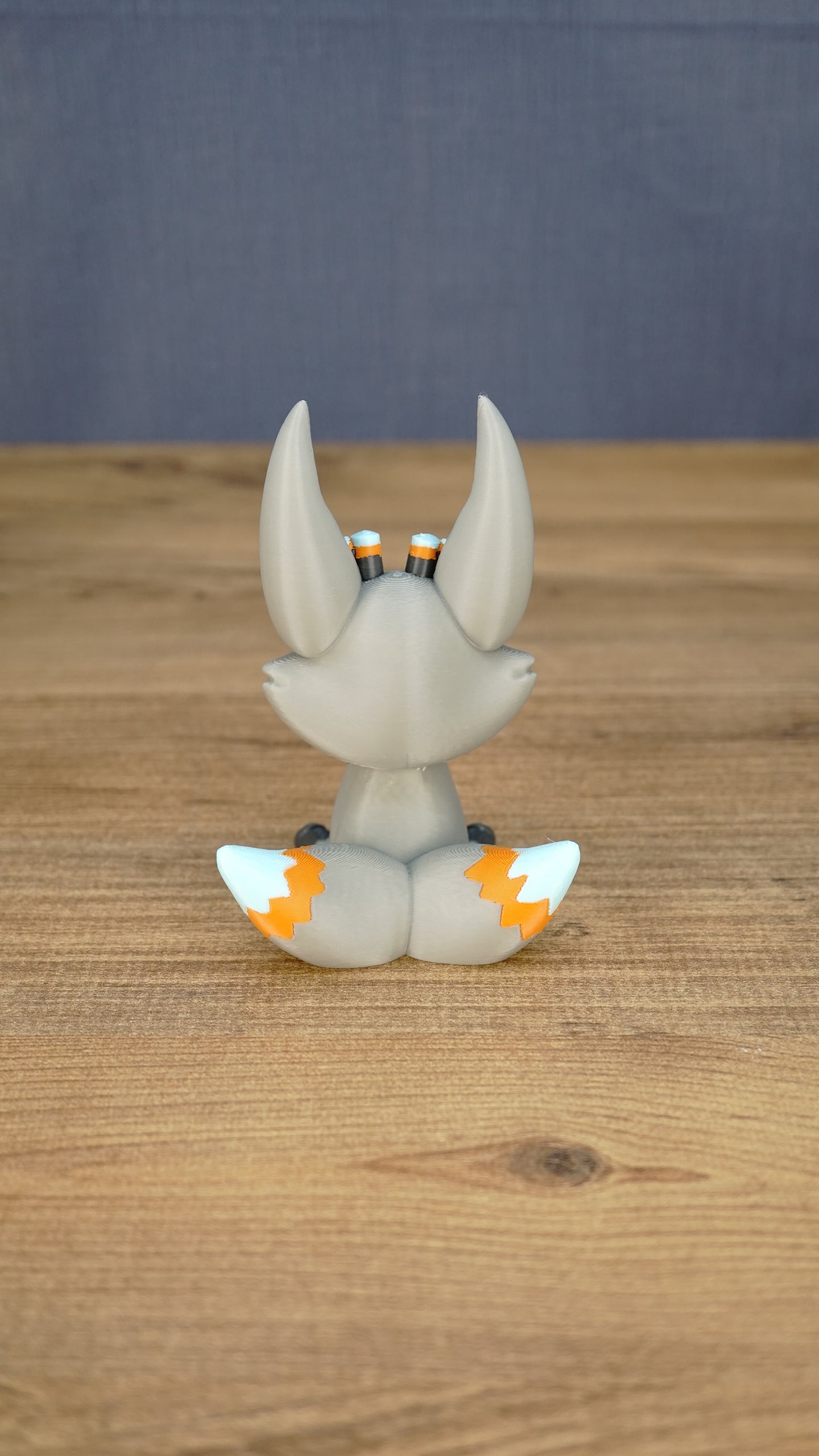 Side view of the Palia Kitsuu Caldera plush figure showing its tall ears, seated pose, gray body, and orange and white tail accents.