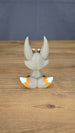 Side view of the Palia Kitsuu Caldera plush figure showing its tall ears, seated pose, gray body, and orange and white tail accents.