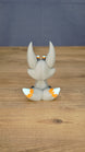 Side view of the Palia Kitsuu Caldera plush figure showing its tall ears, seated pose, gray body, and orange and white tail accents.