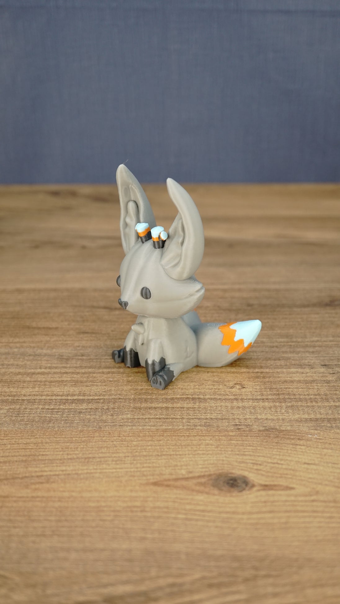 Side view of the Palia Kitsuu Caldera plush figure showing its tall ears, seated pose, gray body, and orange and white tail accents.
