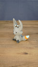 Side view of the Palia Kitsuu Caldera plush figure showing its tall ears, seated pose, gray body, and orange and white tail accents.
