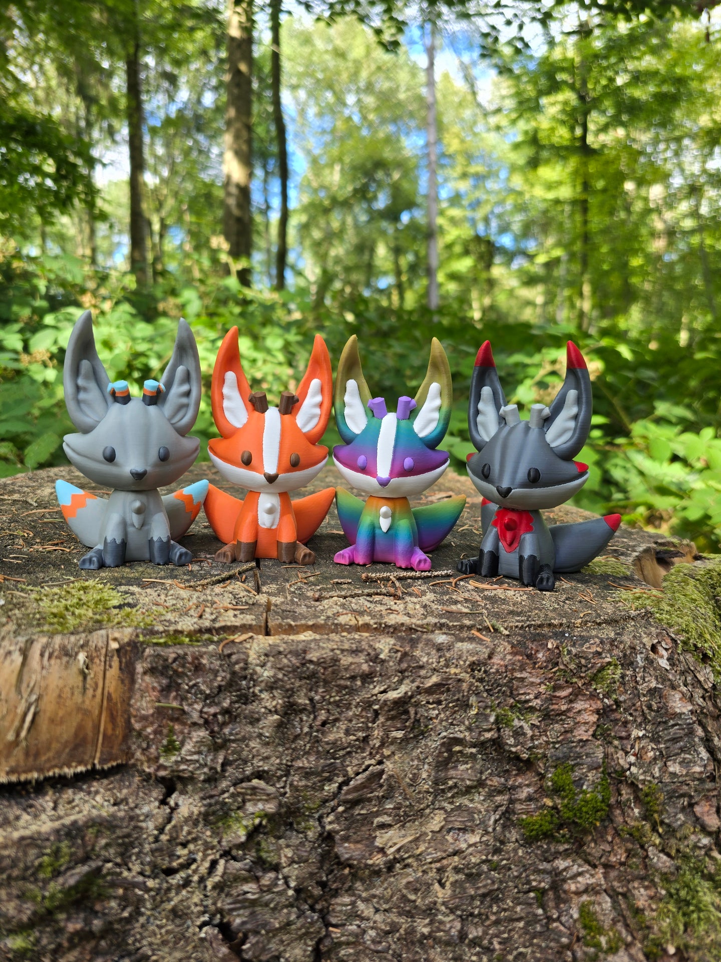 Four 3D printed Palia Kitsuu figures in ancient, orange, rainbow, and obsidian colourways displayed on a tree stump in a forest.
