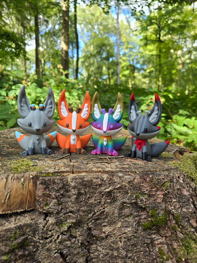 Four 3D printed Palia Kitsuu figures in ancient, orange, rainbow, and obsidian colourways displayed on a tree stump in a forest.