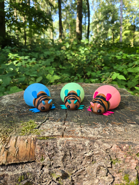 Three colourful Palia Lantern Bug 3D printed plush figures in blue, green, and pink displayed on a tree stump in a forest.