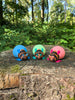 Three colourful Palia Lantern Bug 3D printed plush figures in blue, green, and pink displayed on a tree stump in a forest.
