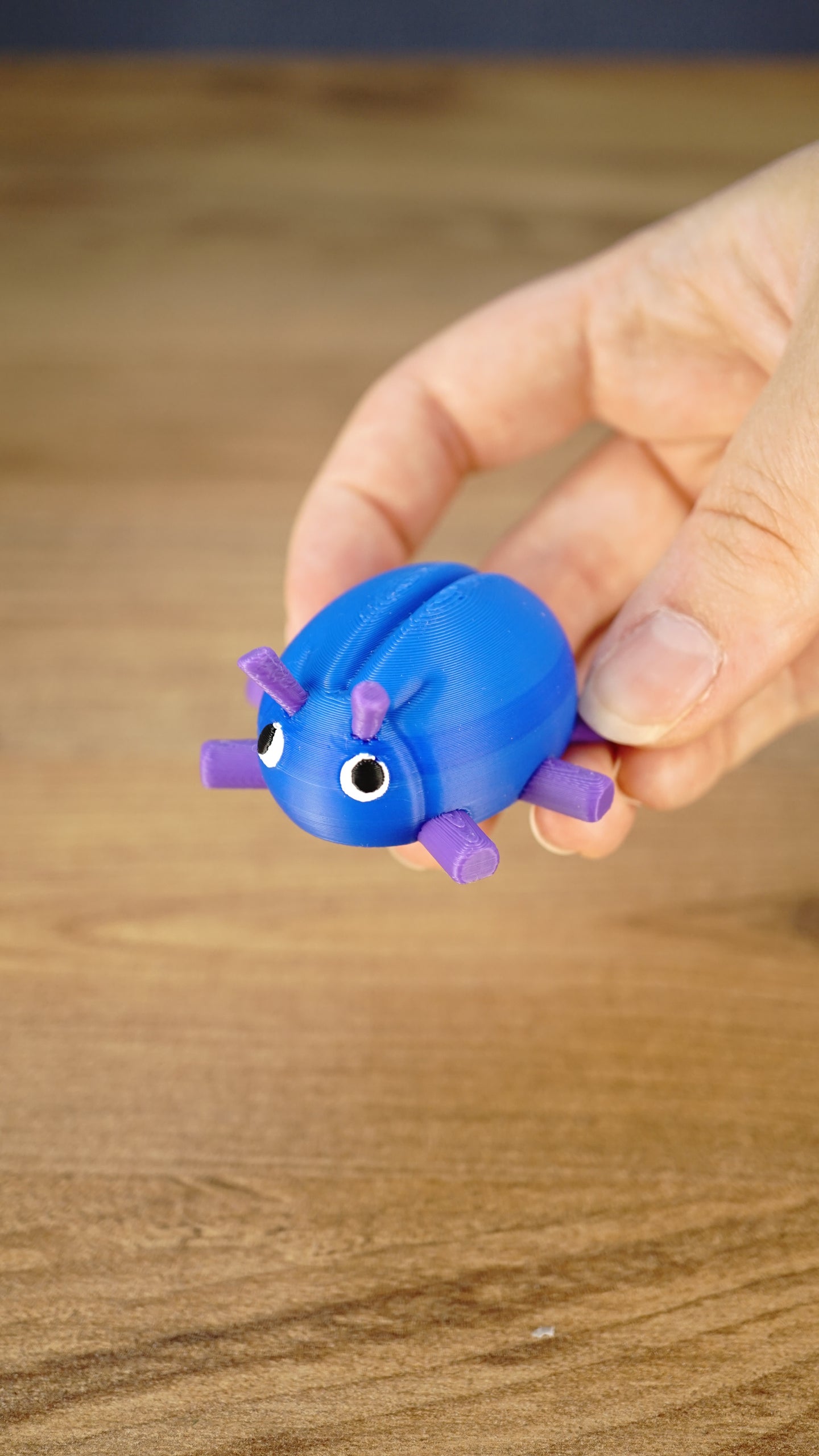 Hand holding the blue and purple Li’l Beetle miniature, showing its rounded shell, six legs, antennae and expressive white eyes.