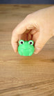 Hand holding the round green Li’l Froggy miniature, highlighting its large expressive eyes, little feet and compact collectible size.