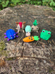 Cute mini 3D printed Palia-inspired plush bundle displayed on a tree stump in the forest, featuring a blue bug, sernuk, snail, sprout, frog, and carrot figure.