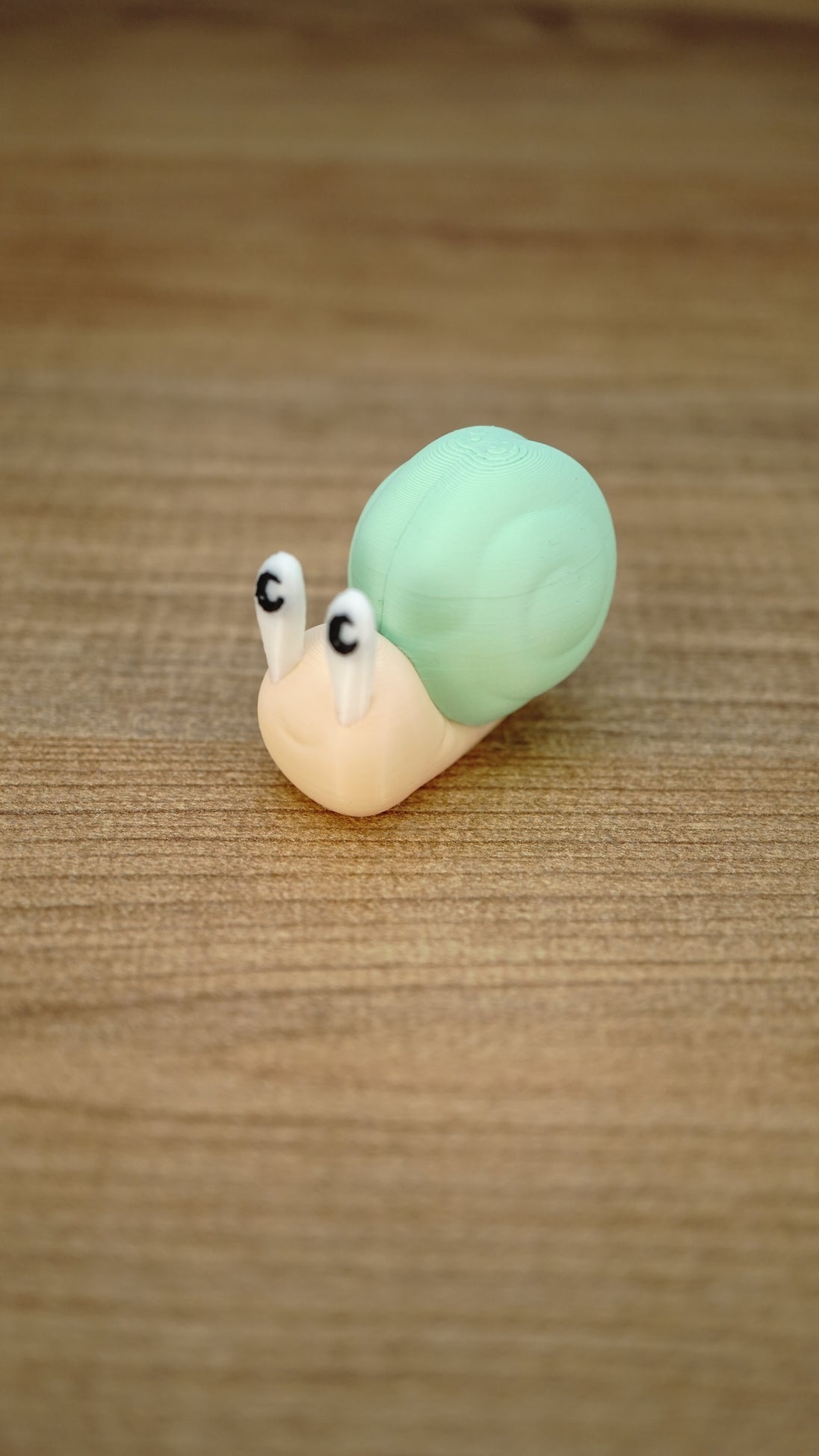 Li’l Snail collectible miniature displayed on a wooden surface with a pale cream body, tall eyes and a soft mint-green shell.