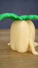 Rear angled view of a Palia Lil Sprout collectible figure showing its smooth cream body, small legs and oversized green leafy sprout design.