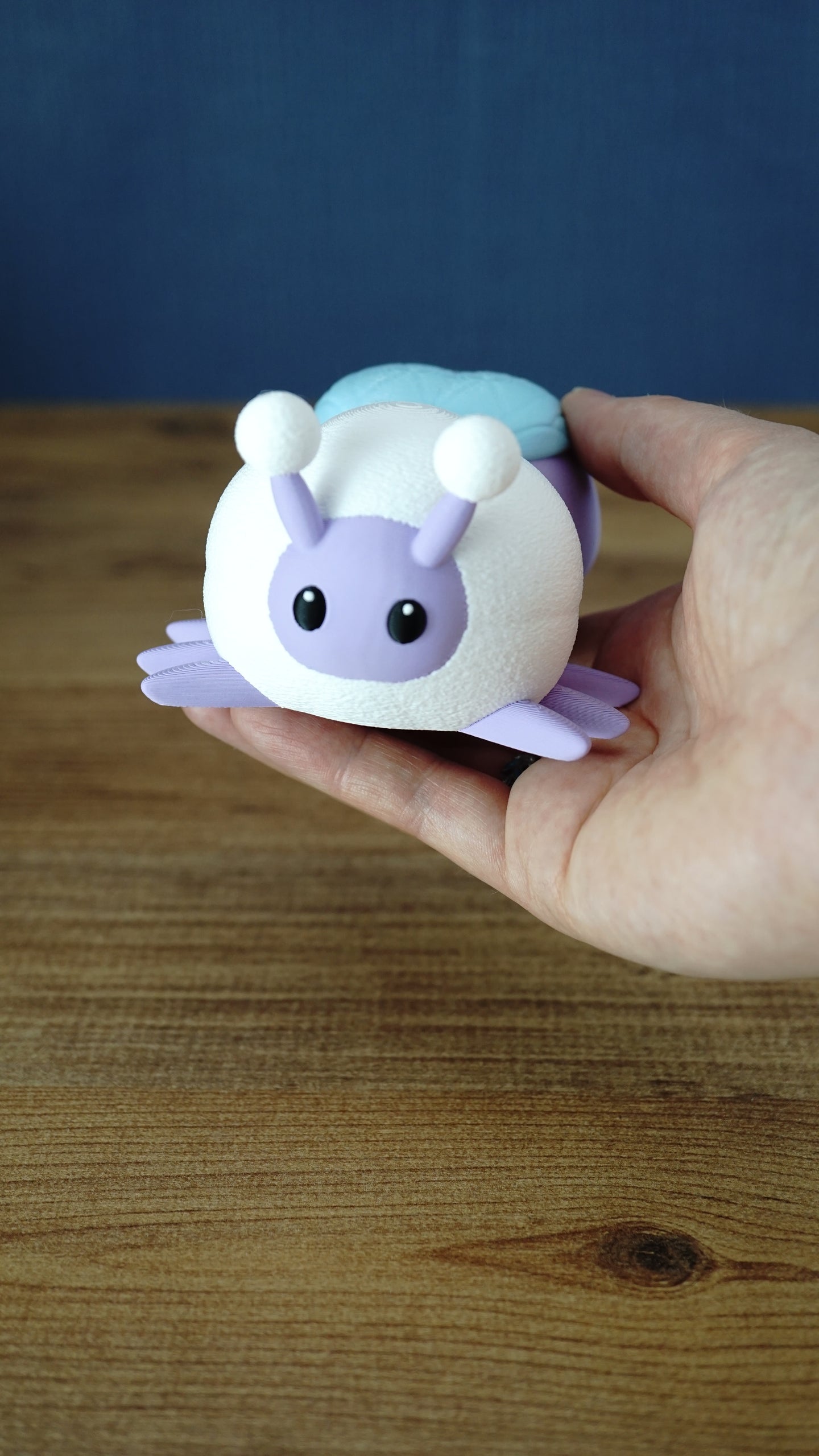 Hand holding a pastel 3D printed Palia Luna Moth plush with lavender face, white fuzzy body, black eyes, and light blue wings.