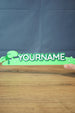 Front view of the Palia New in Town nameplate sign being held to show scale, with bold raised letters and bright green decorative base.