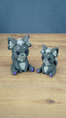 Two 3D printed Palia-inspired Nox the Palcat plush figures in dark grey with pale grey accents, purple eyes, and purple paw pads on a wooden tabletop.