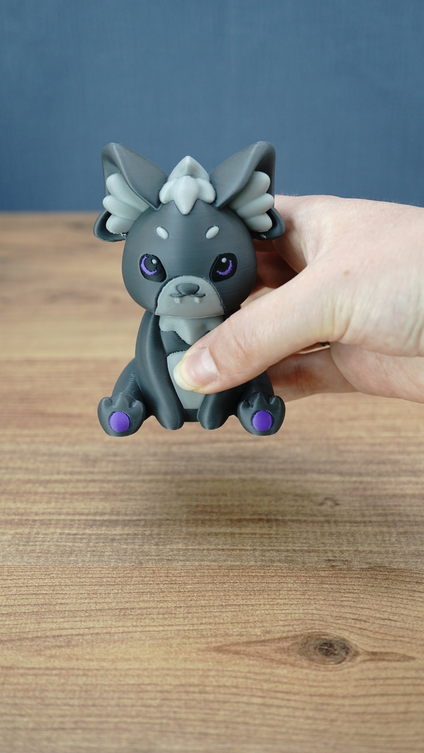 Handheld close-up of a 3D printed Nox the Palcat plush figure with dark grey body, pale grey facial details, purple eyes, and purple paw pads.