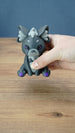 Handheld close-up of a 3D printed Nox the Palcat plush figure with dark grey body, pale grey facial details, purple eyes, and purple paw pads.