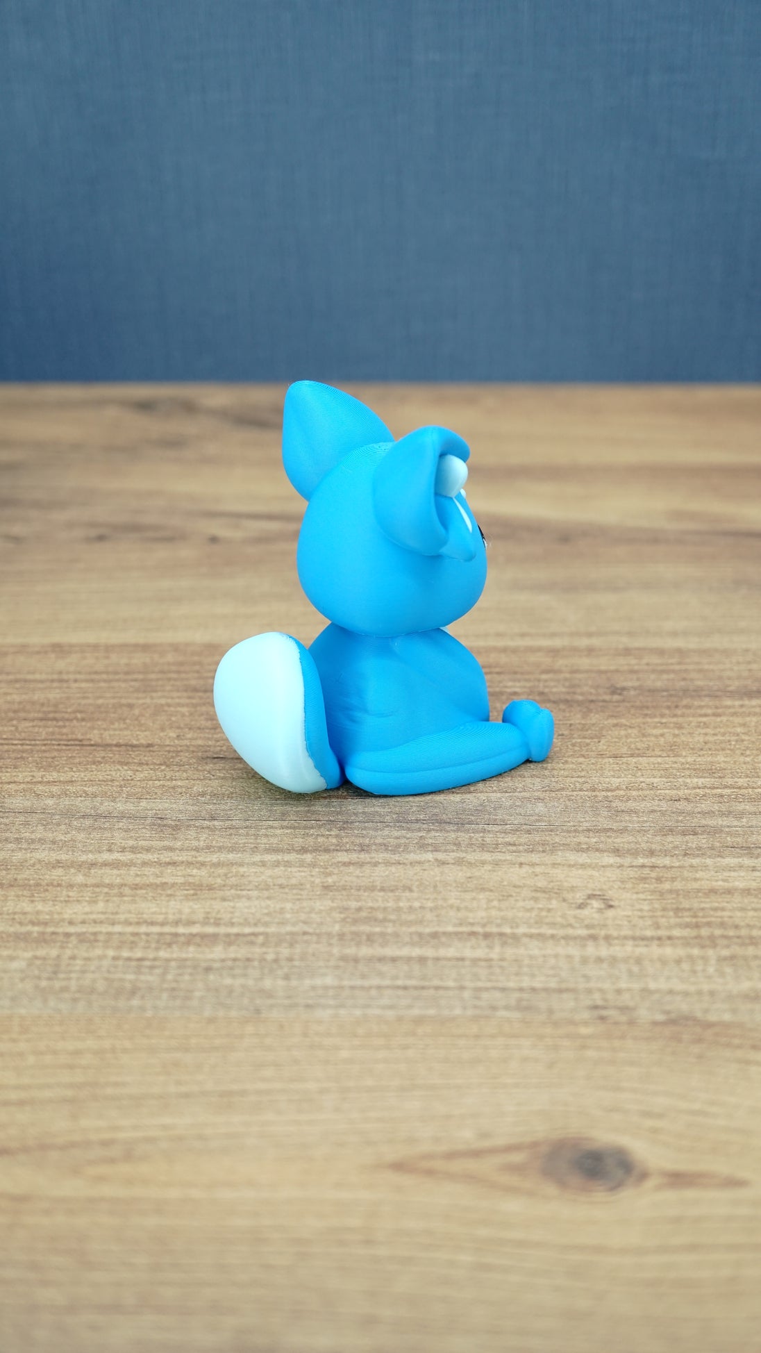 Back side view of a bright blue 3D printed One-in-Ten-Million Palcat plush figure showing its white-tipped curled tail, large ears, and seated body shape.