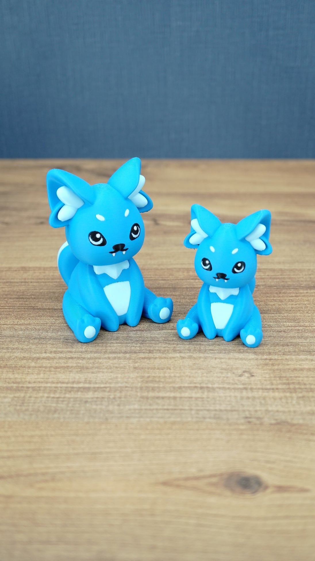 Two bright blue 3D printed Palia-inspired One-in-Ten-Million Palcat plush figures with white accents, black eyes, tiny fangs, and white paw pads on a wooden tabletop.