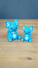 Two bright blue 3D printed Palia-inspired One-in-Ten-Million Palcat plush figures with white accents, black eyes, tiny fangs, and white paw pads on a wooden tabletop.