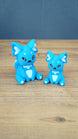 Two bright blue 3D printed Palia-inspired One-in-Ten-Million Palcat plush figures with white accents, black eyes, tiny fangs, and white paw pads on a wooden tabletop.