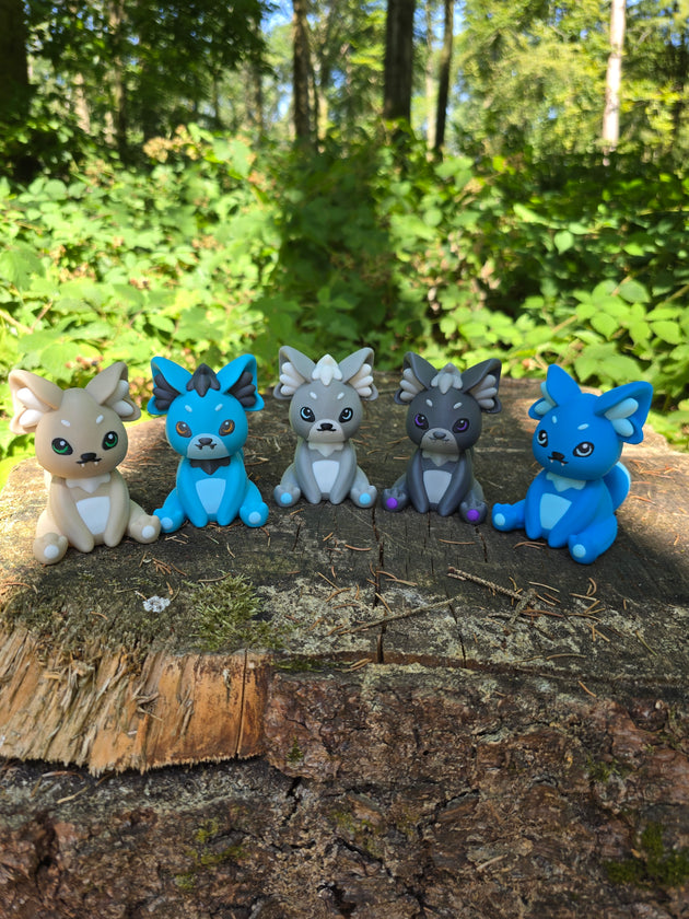 Five 3D printed Palcat plush figures from Palia in cream, blue, grey, dark grey, and bright blue colours displayed on a tree stump in a forest.