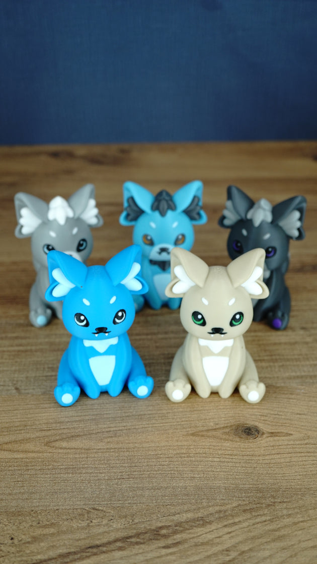 Full collection of 3D printed Palcat plush figures from Palia in blue, cream, grey, dark grey, and turquoise designs on a wooden tabletop.
