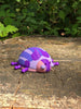 Palia Princess Ladybug collectible displayed outdoors on a woodland tree stump, showing its purple, pink, blue and white design in natural surroundings