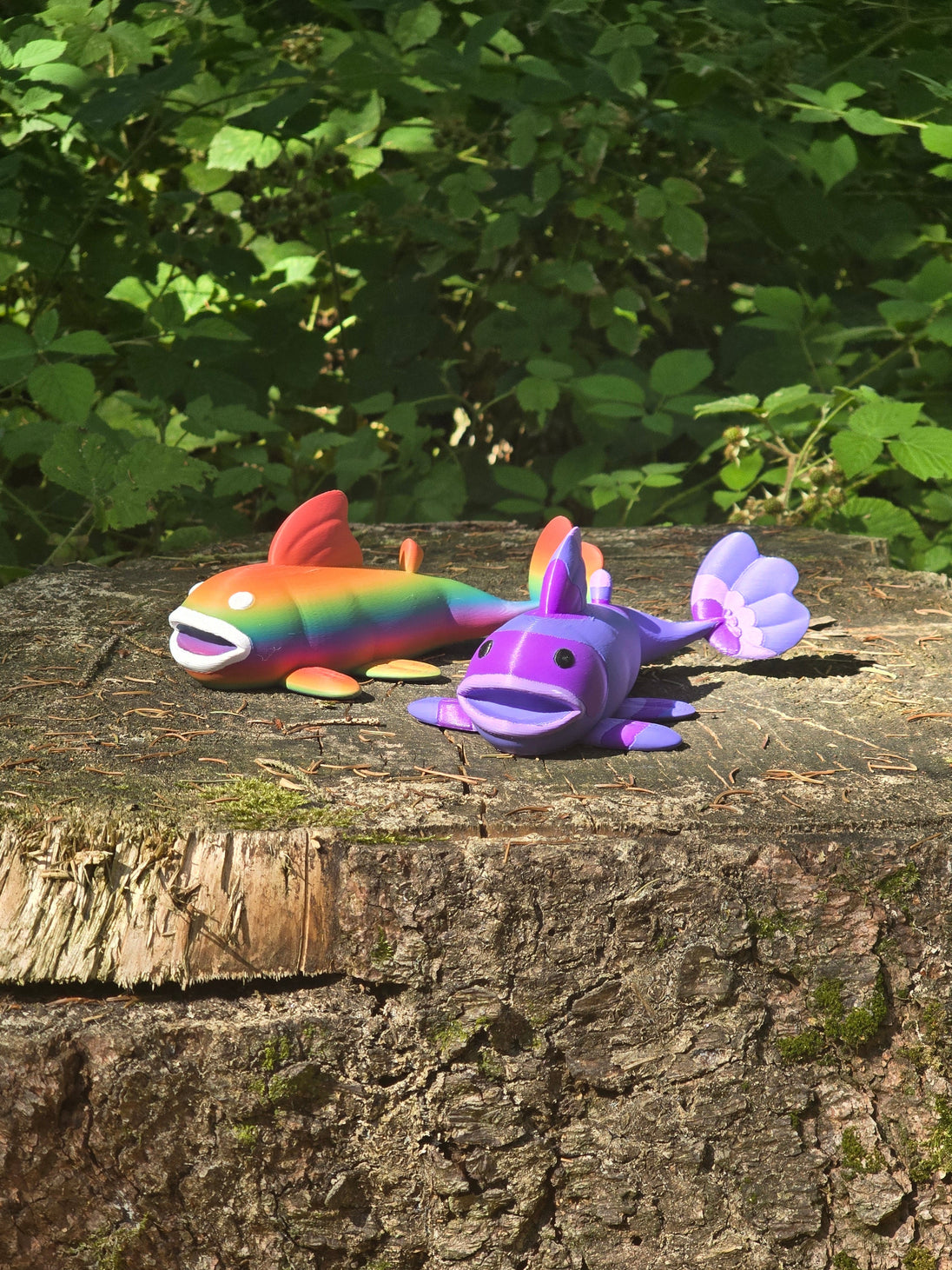 Two 3D printed Palia Prismfish plush figures, one rainbow and one purple, displayed on a mossy tree stump in a forest.