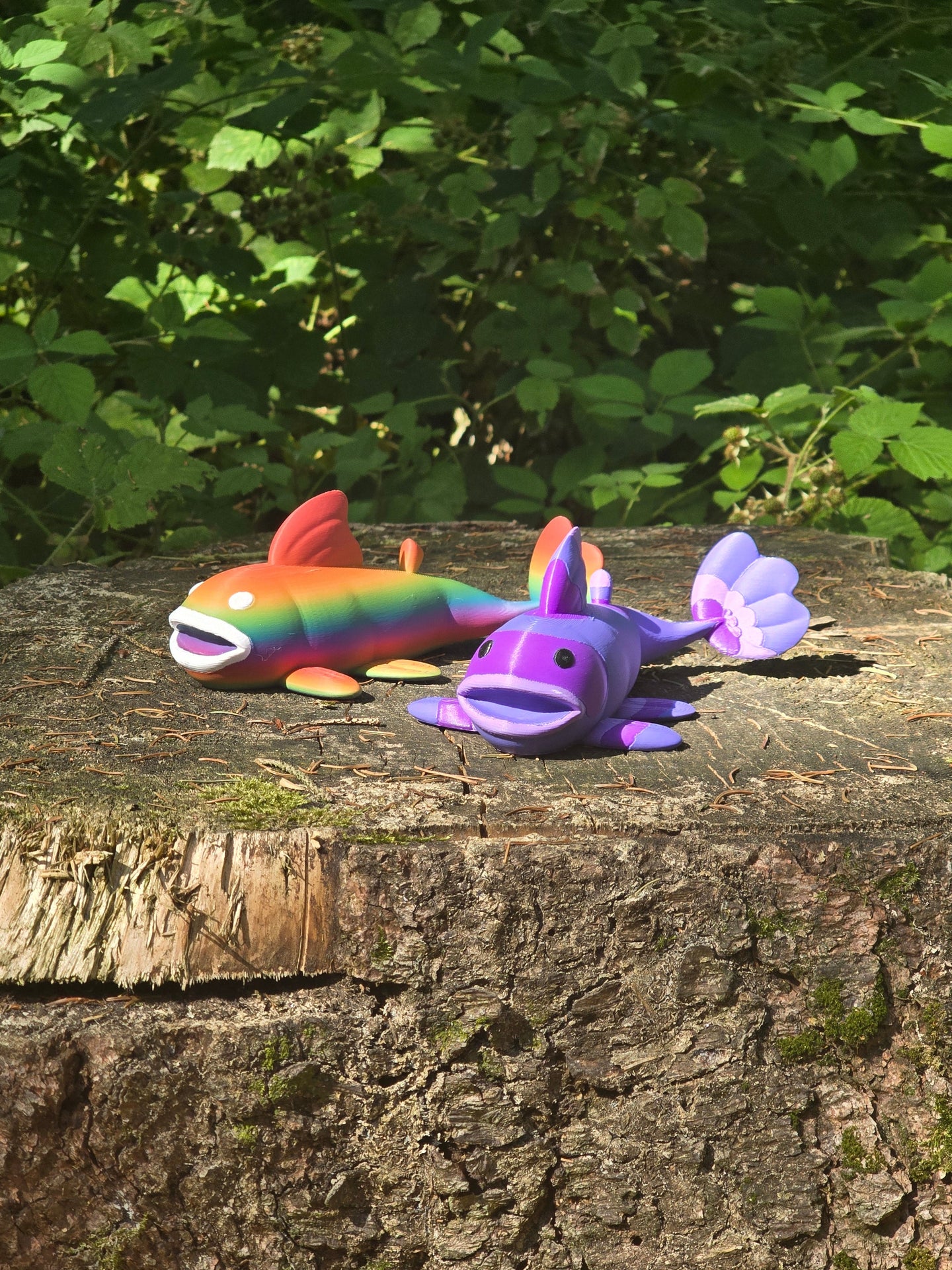 Two 3D printed Palia Prismfish plush figures, one rainbow and one purple, displayed on a mossy tree stump in a forest.