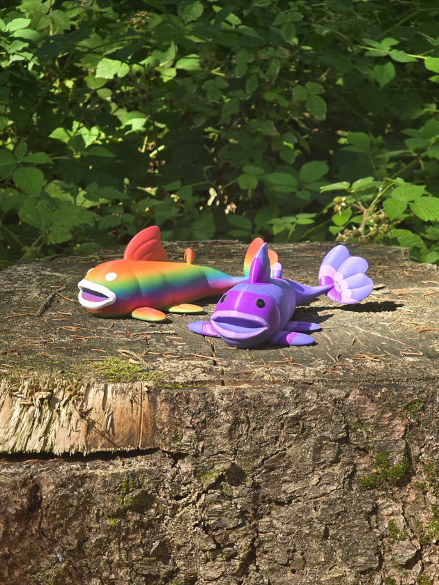 Two 3D printed Palia Prismfish plush figures, one rainbow and one purple, displayed on a mossy tree stump in a forest.