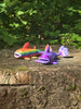 Two 3D printed Palia Prismfish plush figures, one rainbow and one purple, displayed on a mossy tree stump in a forest.