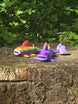 Two 3D printed Palia Prismfish plush figures, one rainbow and one purple, displayed on a mossy tree stump in a forest.