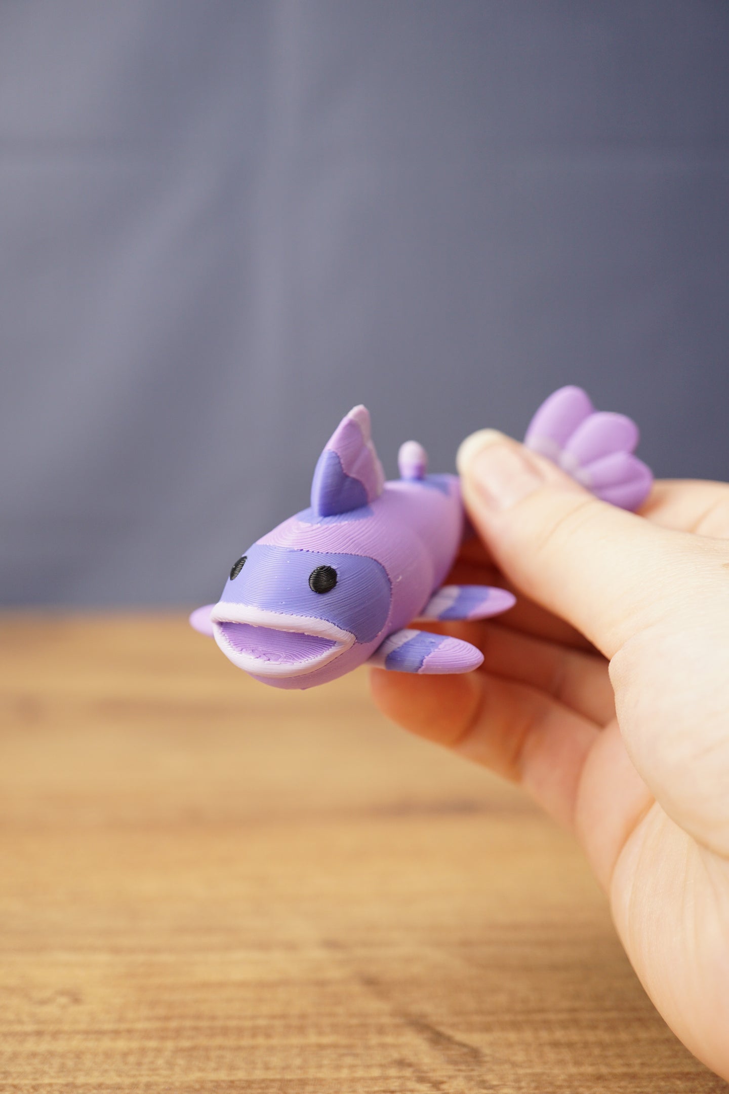 Close-up of a small purple Palia Prismfish 3D printed figure being held, with lavender fins, black eyes, and an open mouth.