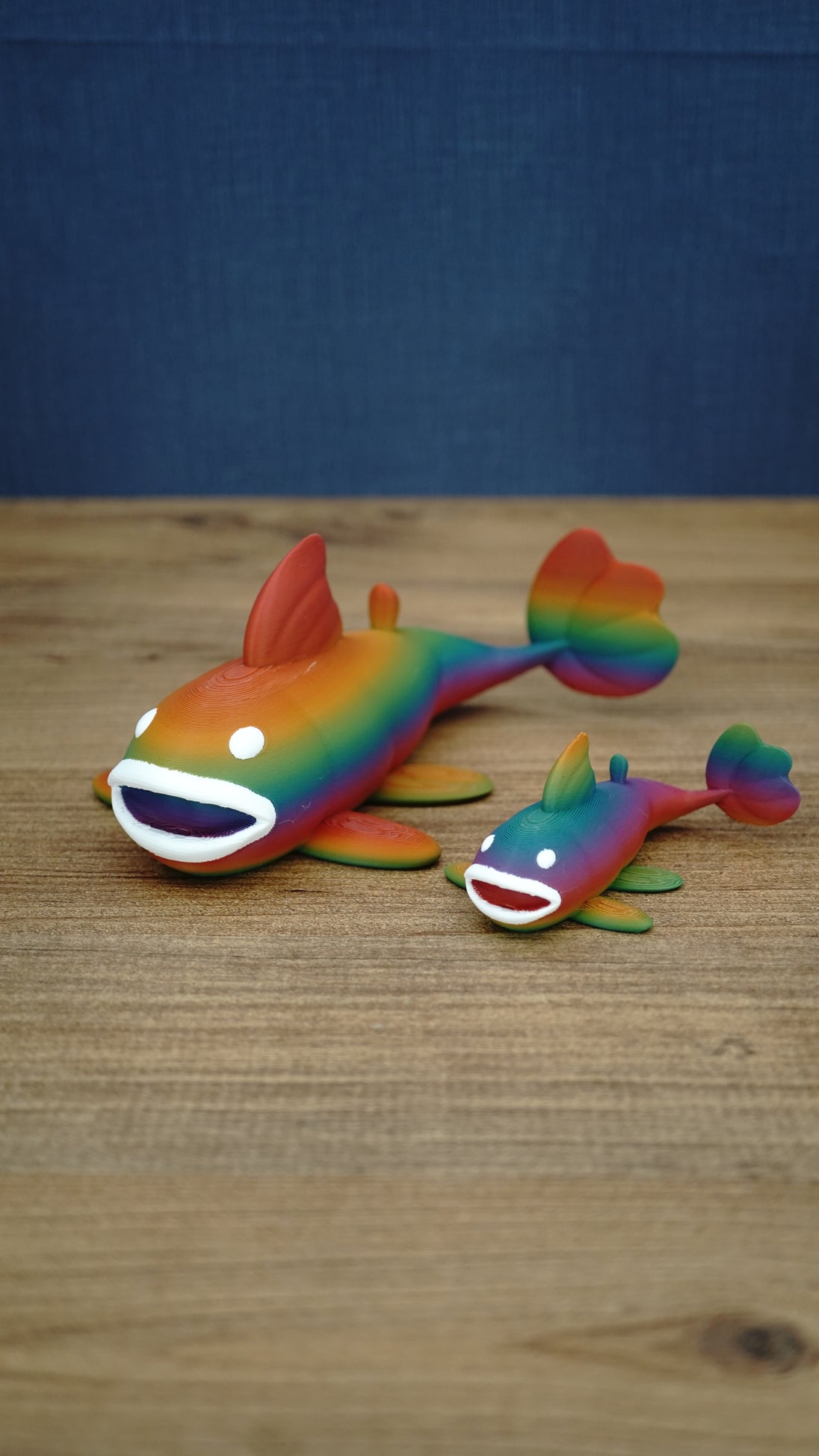 Two rainbow Palia Prismfish 3D printed figures displayed on a wooden tabletop, featuring bright rainbow colouring and white eyes.