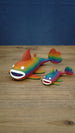 Two rainbow Palia Prismfish 3D printed figures displayed on a wooden tabletop, featuring bright rainbow colouring and white eyes.